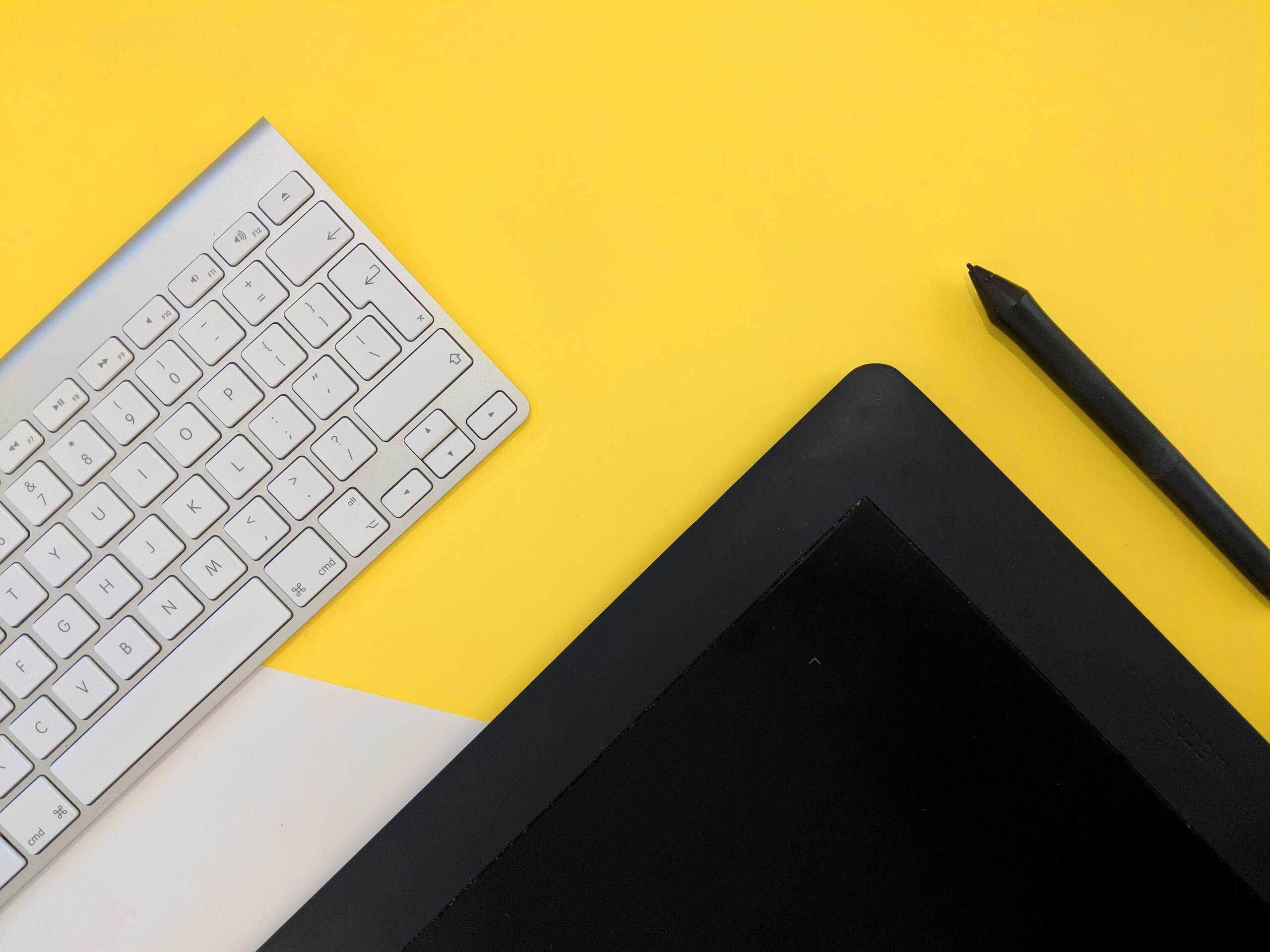 A white Apple keyboard and black iPad sit on a yellow desk or tabletop.